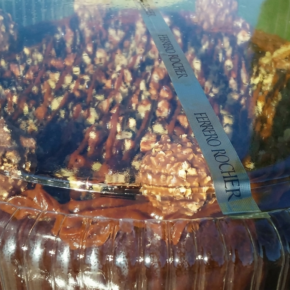 Ferrero Rocher chocolate-covered cake or dessert in a clear plastic container, topped with nuts and chocolate drizzle.