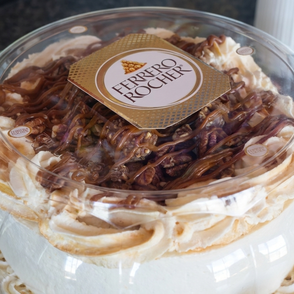 Layered Ferrero Rocher cake topped with chocolate drizzle and hazelnuts in a clear plastic container.